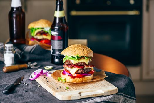 Burger with Two Beef Patties, Cheddar Cheese, Bacon, Iceberg Lettuce, Sliced Tomatoes and Red Onion on Cutting Board. Bottle of Beer and Some Ingredients on Table.