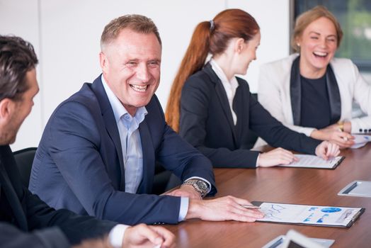 Business people at meeting table discuss documents with reports and diargams showing financial statistics and laughing