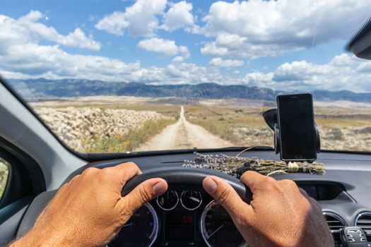 Detail of male driver hands on steering wheel. Driving a car on country road on dramatic countryside lanscape of Pag island, Croatia. View from the cabin trough the windshield.