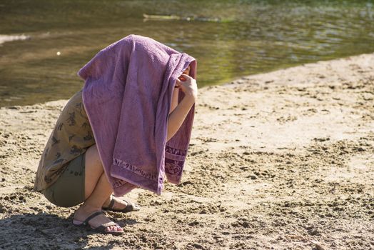 The girl hid her face from the bright sun under a towel.The girl is sitting on the beach with a towel on her head.