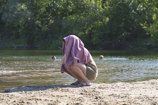 The girl hid her face from the bright sun under a towel.The girl is sitting on the beach with a towel on her head.