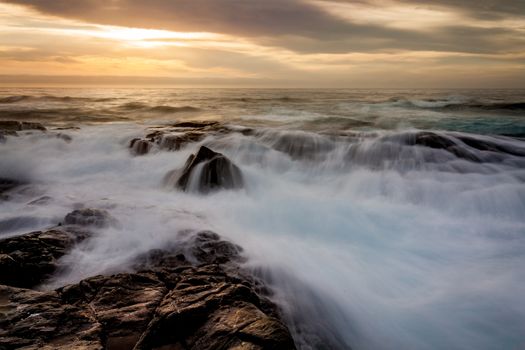 Mystical light filters through clouds as waves cascade over a rocky craggy seashore