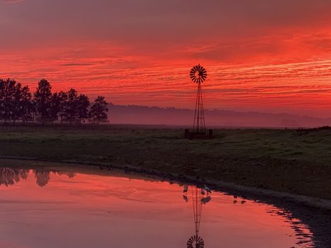 Light morning fog, windmill, pond and a stunning vibrant red sunrise sky in rural countryside farmlands of Australia