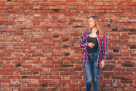 Portrait of young woman in checkered shirt and blue jeans standing against red brick wall