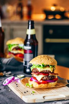 Cheeseburger with Two Beef Patties, Cheddar Cheese, Bacon, Iceberg Lettuce, Sliced Tomatoes and Red Onion. Bottle of Beer and Some Ingredients on Table.
