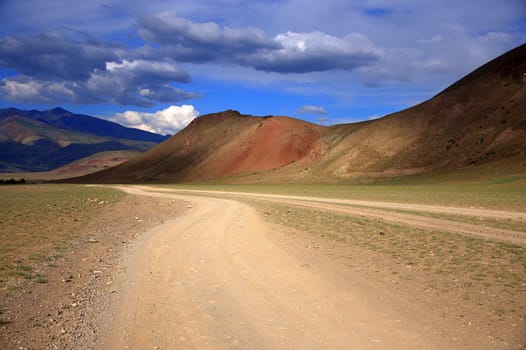 Dirt road winds at the foot of the red mountains under the thunderclouds in the sky. Altai, Siberia, Russia