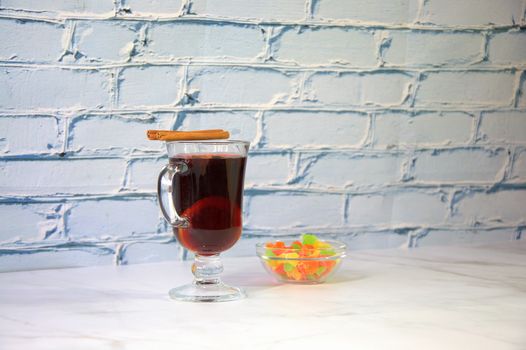 A glass of hot mulled wine with lemon and cinnamon sticks stands on a table near a bowl of dried fruit.