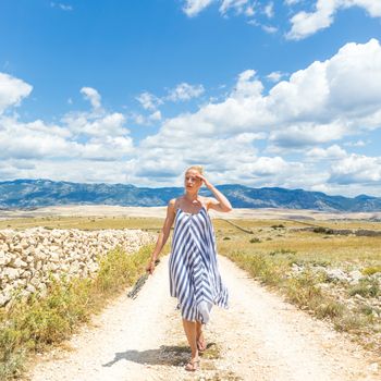 Caucasian young woman in summer dress holding bouquet of lavender flowers while walking outdoor through dry rocky Mediterranean Croatian coast lanscape on Pag island in summertime.