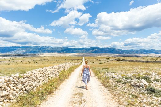 Caucasian young woman in summer dress holding bouquet of lavender flowers while walking outdoor through dry rocky Mediterranean Croatian coast lanscape on Pag island in summertime.