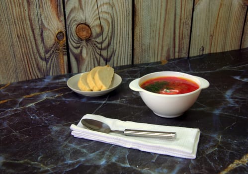 Traditional Russian soup Borsch with sour cream and herbs, stands on a table next to pieces of bread on a plate and a napkin with a spoon.