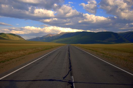 A direct asphalt road going beyond the horizon rests on mountain ranges. Kurai steppe, Altai, Russia.