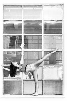 Fit sporty active girl in fashion sportswear doing yoga fitness exercise in front of big industrial window frame. colorful reflections in window glass. Urban style yoga. Black and white.