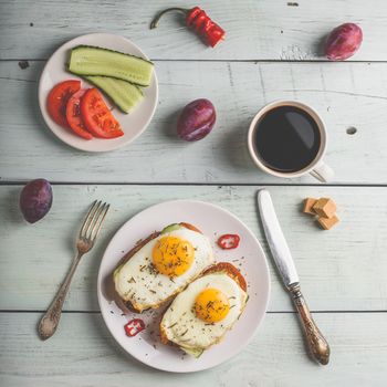 Breakfast toasts with vegetables and fried egg on white plate, cup of coffee and some fruits over wooden background. Healthy food concept. View from above.