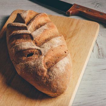 Homemade rye loaf on cutting board with knife over light wooden background.