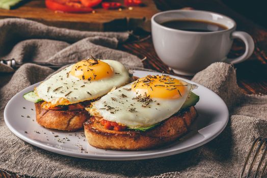 Italian toasts with vegetables and fried eggs on white plate and cup of coffee over grey rough cloth.