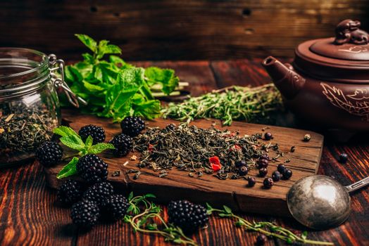 Heap of Dry Green Tea and Fresh Blackberries on Wooden Cutting Board. Bundles of Mint and Thyme Leaves. Clay Kettle.