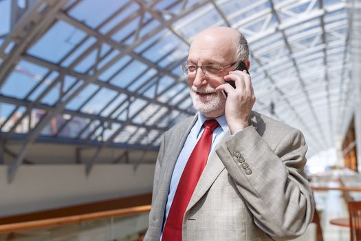 Senior business man dressed in suit and eyeglasses is talking on the mobile phone and smiling while sitting in modern building