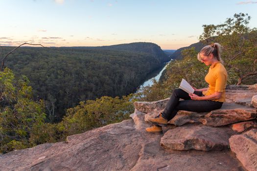A woman sitting on a rock reading a book out in nature cliff edge with river views
