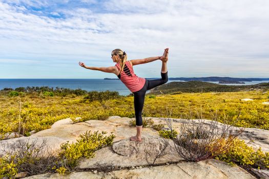 Female in outdoor coastal setting with wildflowers blooming doing yoga asanas. Natarajasana, or Lord of the Dance Pose