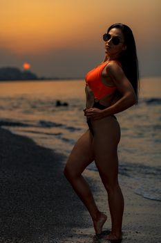 Fitness girl posing 
in the shore of the beach with a beautiful black and orange bikini and sunglasses