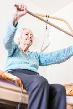 Elderly 96 years old woman exercising with a stick sitting on her bad. Geriatric health care home assisted support for older people concept. Care for the elderly.