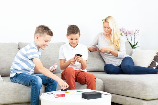 Two kids playing card game on living room sofa at home supevised by their mother. Spending quality leisure time with children and family concept. Cards are generic and debranded.