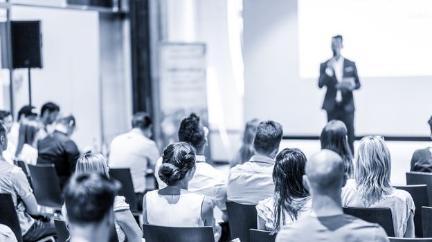 Business and entrepreneurship symposium. Speaker giving a talk at business meeting. Audience in conference hall. Rear view of unrecognized participant in audience.