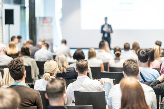 Business and entrepreneurship symposium. Speaker giving a talk at business meeting. Audience in conference hall. Rear view of unrecognized participant in audience.