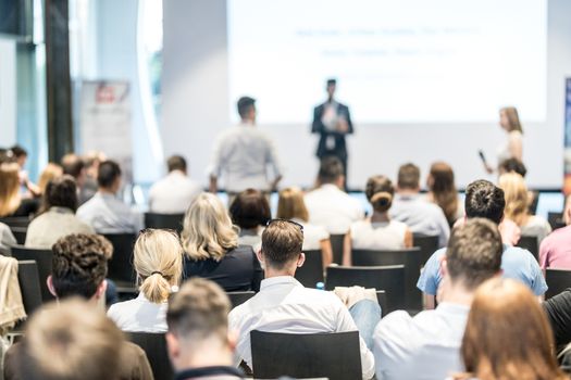 Business and entrepreneurship symposium. Speaker giving a talk at business meeting. Audience in conference hall. Rear view of unrecognized participant in audience.