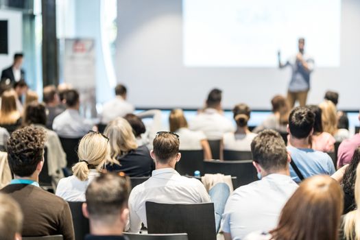 Business and entrepreneurship symposium. Speaker giving a talk at business meeting. Audience in conference hall. Rear view of unrecognized participant in audience.