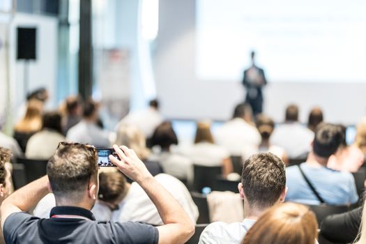 Business and entrepreneurship symposium. Speaker giving a talk at business meeting. Audience in conference hall. Rear view of unrecognized participant in audience taking photo of presentation.