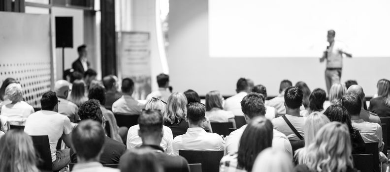 Business and entrepreneurship symposium. Speaker giving a talk at business meeting. Audience in conference hall. Rear view of unrecognized participant in audience.