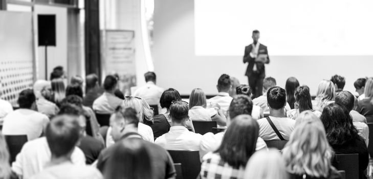 Business and entrepreneurship symposium. Speaker giving a talk at business meeting. Audience in conference hall. Rear view of unrecognized participant in audience.