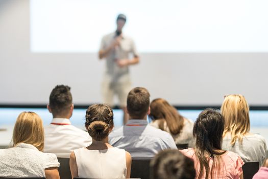 Business and entrepreneurship symposium. Speaker giving a talk at business meeting. Audience in conference hall. Rear view of unrecognized participant in audience.