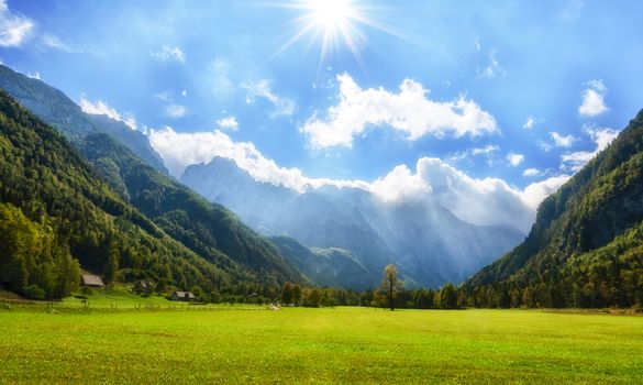 Alpine valley Logarska dolina, Slovenia, green pasture with forest and mountains in background, clouds on blue sky and sunstars for effect, outdoor tourism and travelling, warm and bright scene, panorama