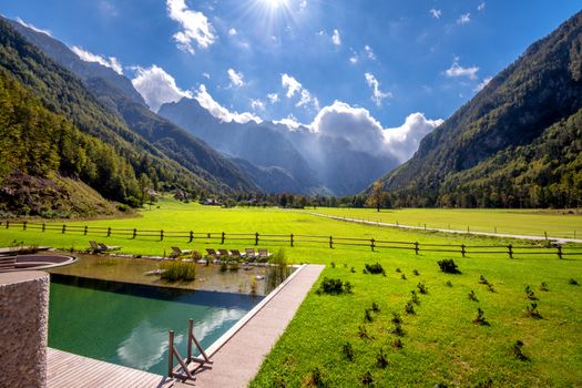 Panoramic view in Logarska valley, Slovenia, green meadows with forest and high mountains in background, natural pond, swimming pool with sunchairs in front