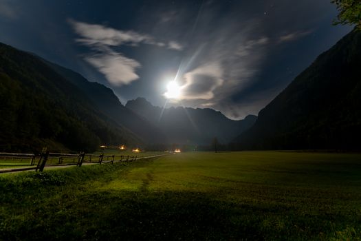 Alpine valley at night, landscape lit by full moon, stars visible, farmhouse with illuminated windows, mistyc and dreamy
