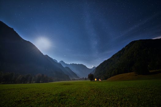 Alpine valley at night, landscape lit by full moon, stars visible, farmhouse with illuminated windows, mistyc and dreamy