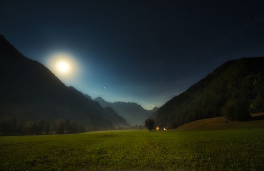 Alpine valley at night, landscape lit by full moon, stars visible, farmhouse with illuminated windows, mistyc and dreamy