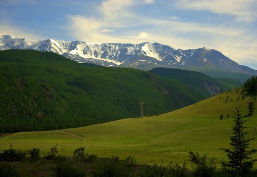 A fertile pasture in a valley near the slopes of a green hill and snow caps of mountain peaks in the background. North Kurai Range, Altai, Siberia, Russia.