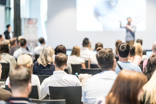 Business and entrepreneurship symposium. Speaker giving a talk at business meeting. Audience in conference hall. Rear view of unrecognized participant in audience.