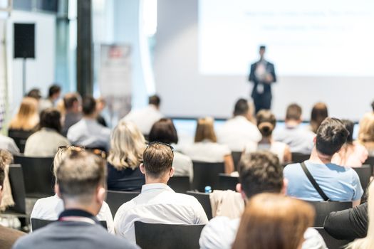 Business and entrepreneurship symposium. Speaker giving a talk at business meeting. Audience in conference hall. Rear view of unrecognized participant in audience.