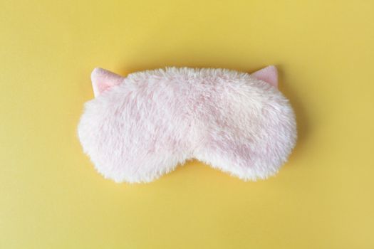 Pastel pink fluffy fur sleep mask with small ears on pastel yellow paper background. Top view, flat lay. Concept of vivid dreams. Accessories for girls and young women.