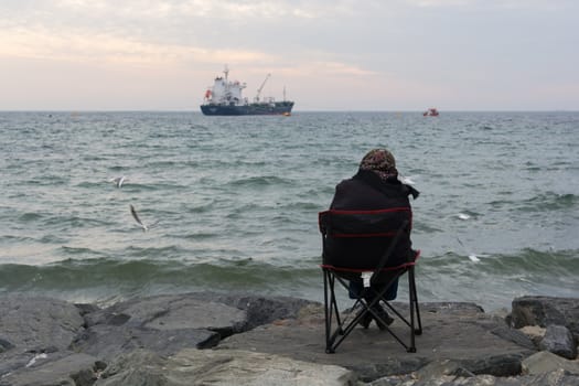 old woman is sitting by the beach. away, looking at ships and birds.considerate