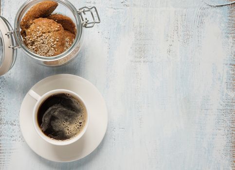 Black coffee with crema in white porcelain cup and oatmeal cookie in a glass jar on a light wooden background with space for text, top view