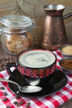 Black and red cup of coffee, checkered napkin, oatmeal cookies in a glass jar and coffee maker on a dark wooden background