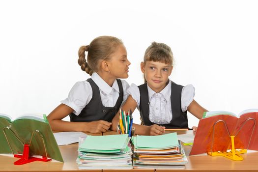 Girls are sitting at a school desk, one whispers to the other something in the ear