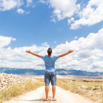 Rear view of casual sporty man standing on a dirt country road rising hands up to the clouds on a blue summer sky. Freedom and travel adventure concept. Copy space.