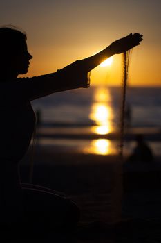 Silhouette of a young girl pouring sand from a beach against a yellow sky and sea at sunset