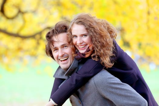 Couple in autumn park having fun, smiling man carrying woman piggyback outdoor